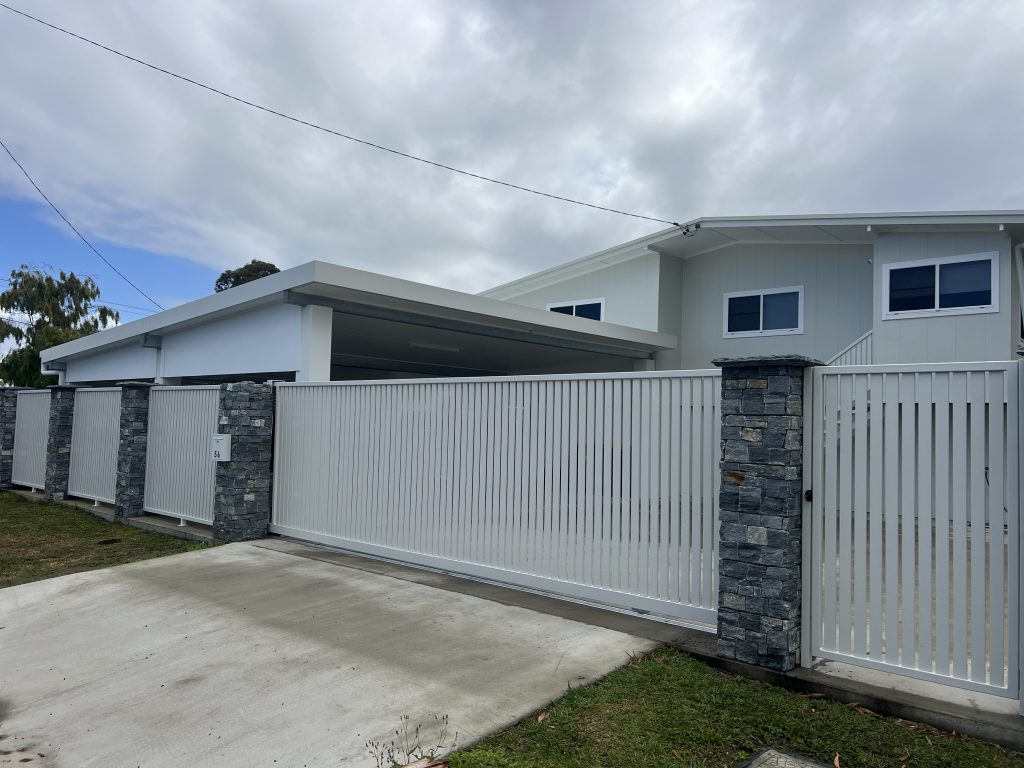 Home with a white slatted sliding gate, stone-clad pillars and concrete driveway