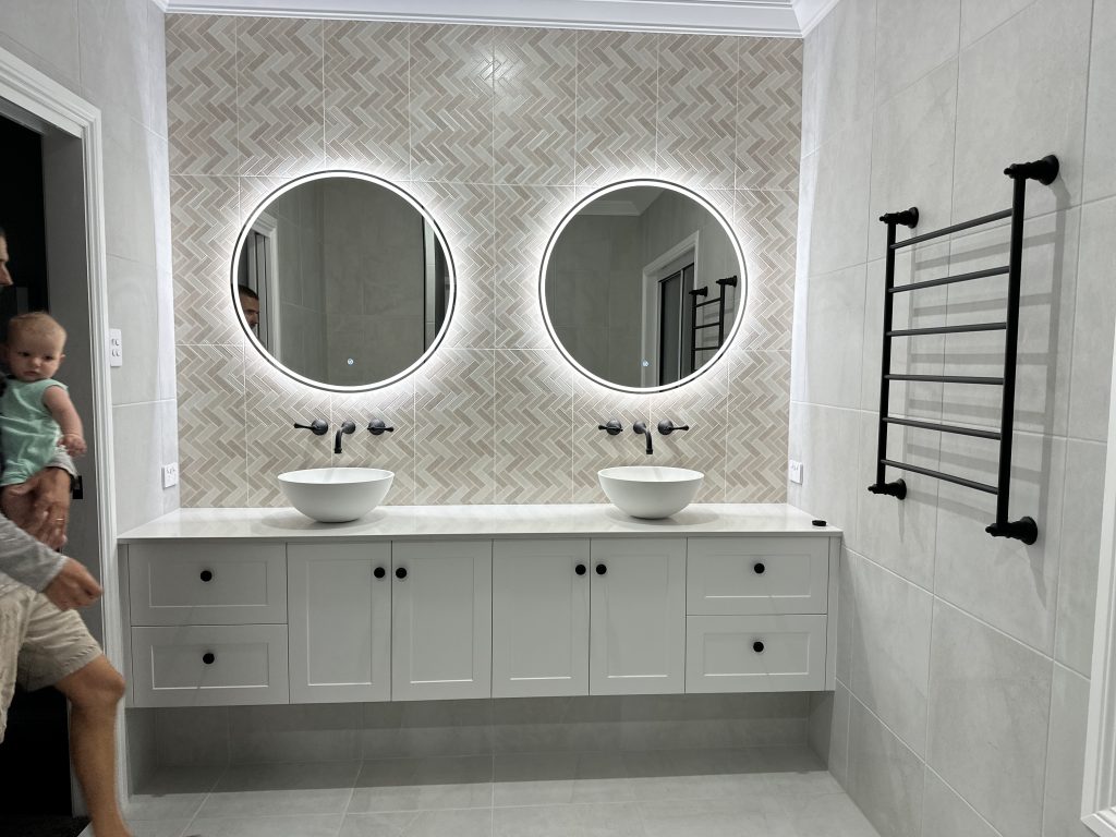 Double vanity with herringbone feature tiles, round backlit mirrors and black towel rail