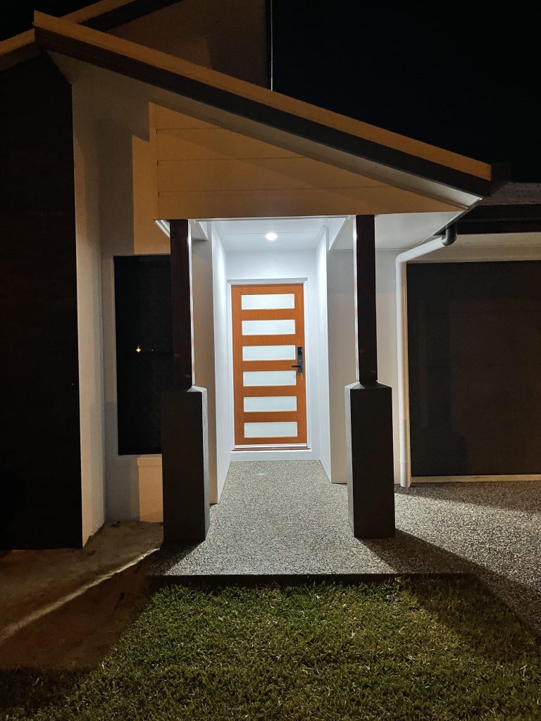 Lit front entry at night with a timber-panelled door and pebble path